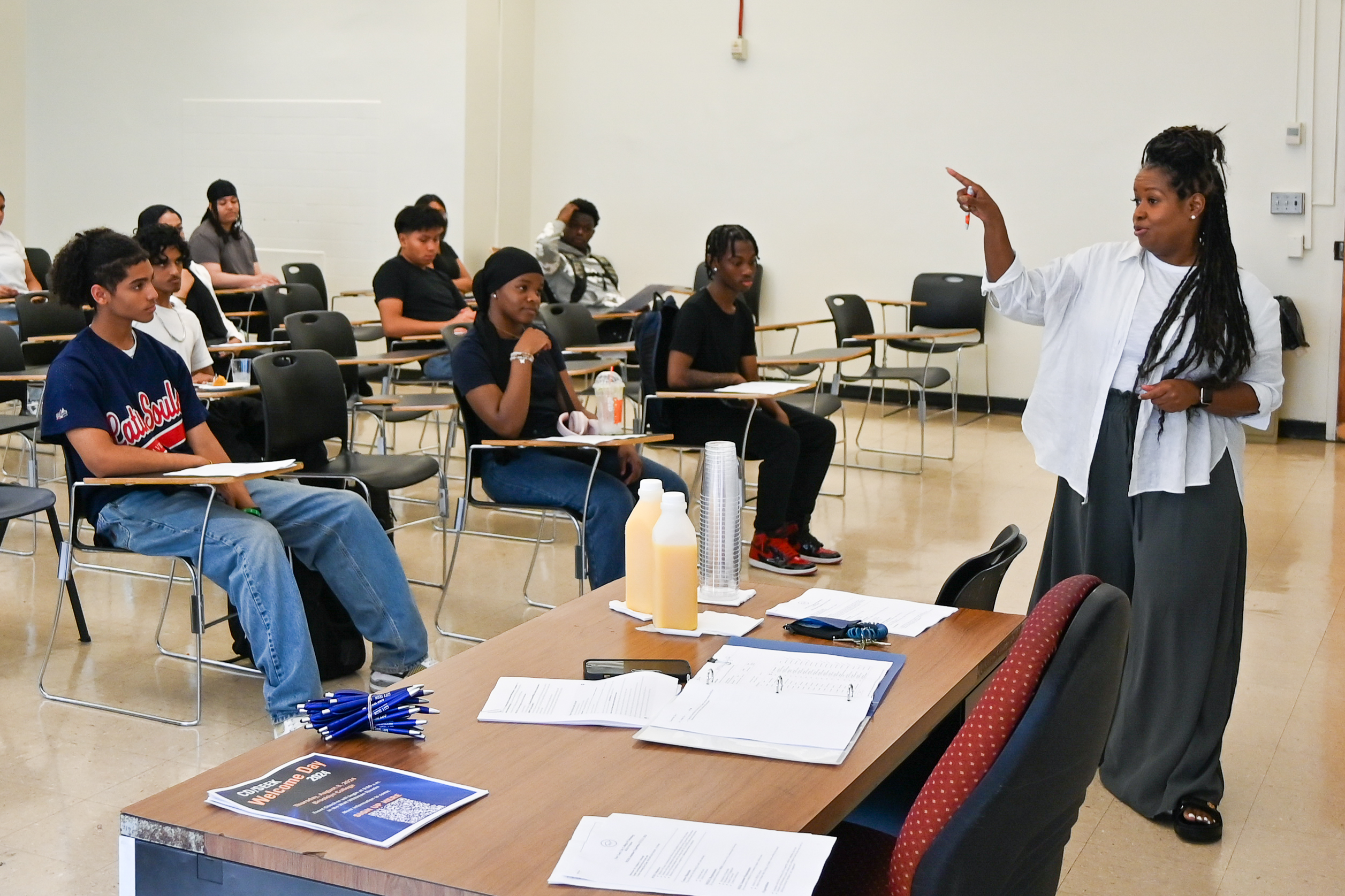 A group of students sitting at desks in a classroom listening to a lecturer speak in the front of the room.