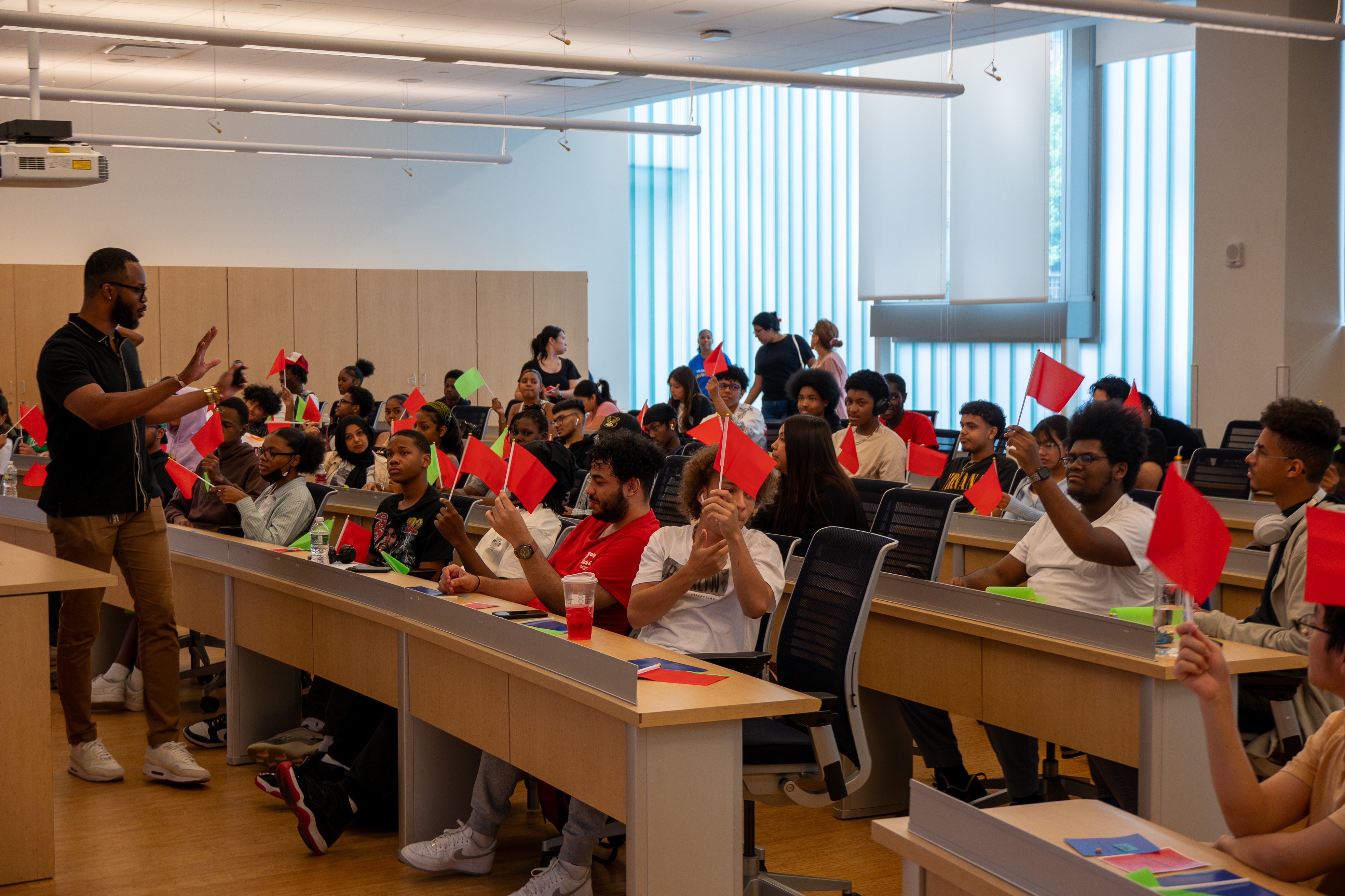 A large group of students sitting in a lecture hall holding up red or green flags as a professor in the front is giving a lecture.