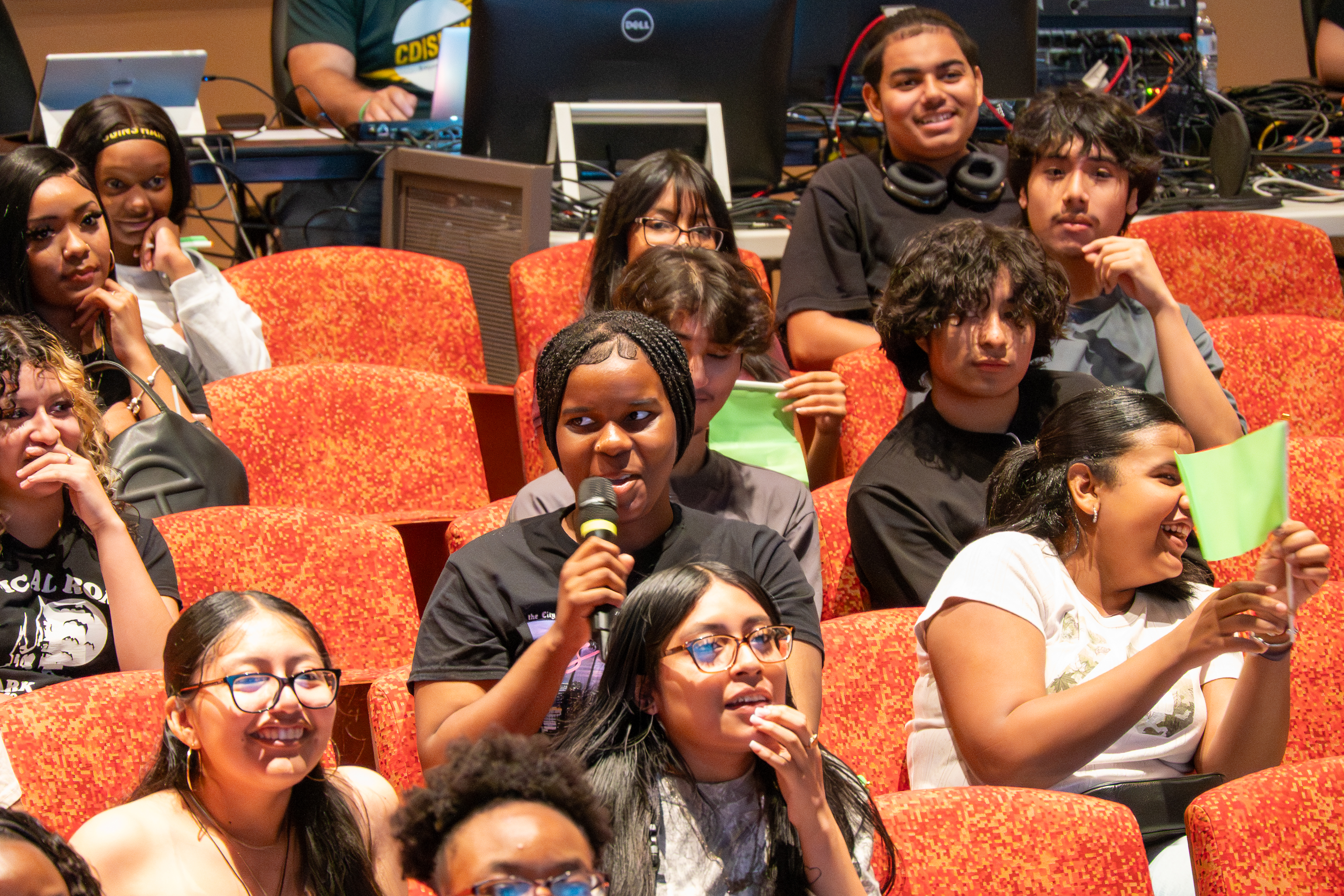 A group of students sitting in the audience of a theater. One student is holding a microphone and speaking into it.